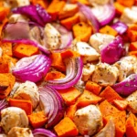 Vibrant close-up of Sheet Pan Chicken with Sweet Potatoes, red onions, and herbs on a rustic baking sheet.