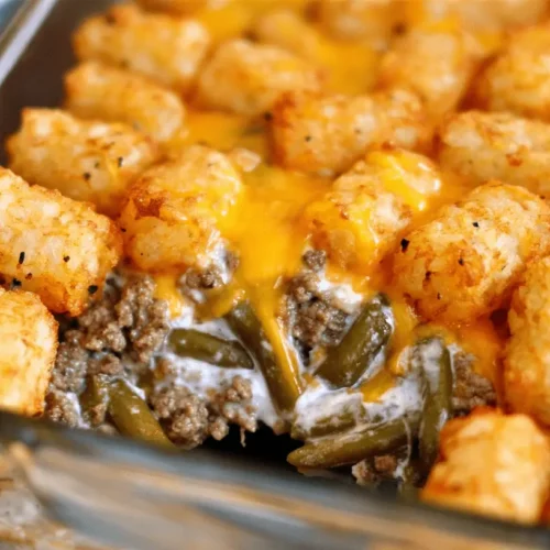 Close-up of a golden brown Tater Tot Casserole bubbling with melted cheese, ground meat, and green beans in a clear glass dish.