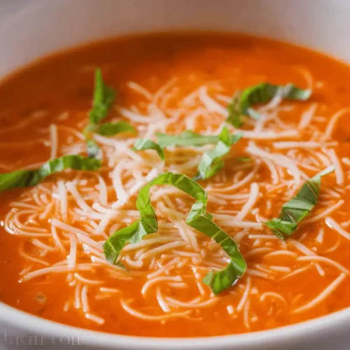 A vibrant close-up of creamy tomato soup topped with fresh basil and white strands, served in a white ceramic bowl.