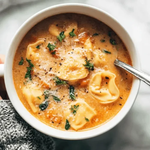 A comforting close-up of a person holding a white bowl of creamy tomato tortellini soup, garnished with cheese and parsley, on a marble surface.