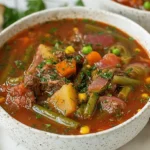 A close-up of a rustic bowl brimming with hearty vegetable beef soup, garnished with fresh herbs and served with crusty bread.