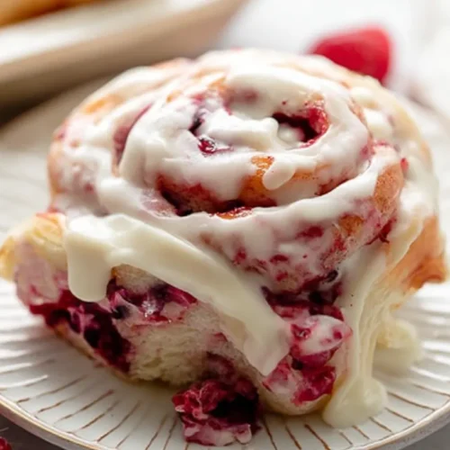 Close-up of a freshly baked Raspberry Swirl Sweet Roll with a generous white glaze and fresh raspberries on a ceramic plate.