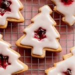 Close-up of festive Christmas tree-shaped Iced Cherry Almond Linzer Cookies with vibrant cherry jam centers on a cooling rack.