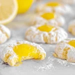 Close-up of freshly baked Lemon Curd Thumbprints dusted with powdered sugar, one broken to reveal the luscious lemon filling, with fresh lemons and a daisy.
