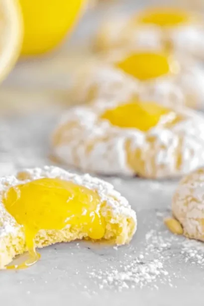 Close-up of freshly baked Lemon Curd Thumbprints dusted with powdered sugar, one broken to reveal the luscious lemon filling, with fresh lemons and a daisy.