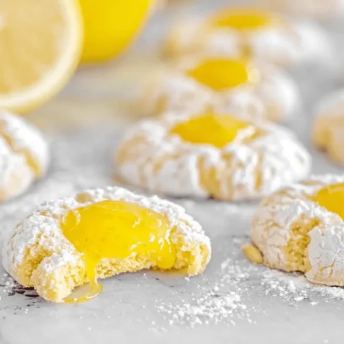 Close-up of freshly baked Lemon Curd Thumbprints dusted with powdered sugar, one broken to reveal the luscious lemon filling, with fresh lemons and a daisy.